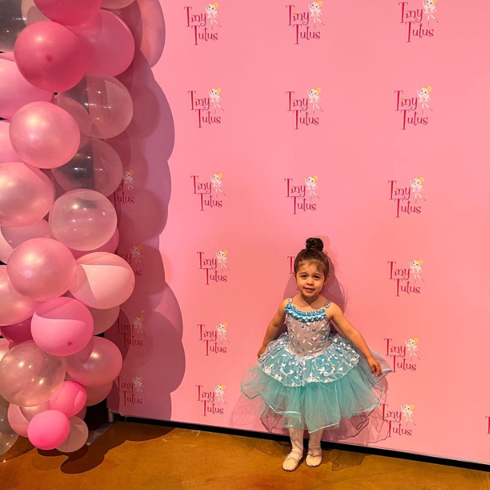 3 year old ballerina in a blue tutu posing in front of a Tiny Tutus ballet media wall at a dance recital. 