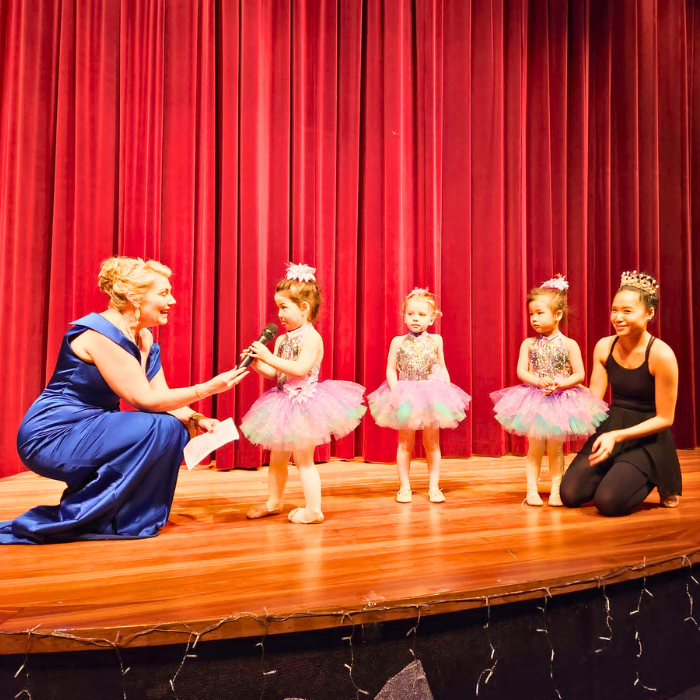 Three young ballerinas in tutus on stage with their teacher and event host at a Tiny Tutus recital. One ballerina is speaking into microphone. 