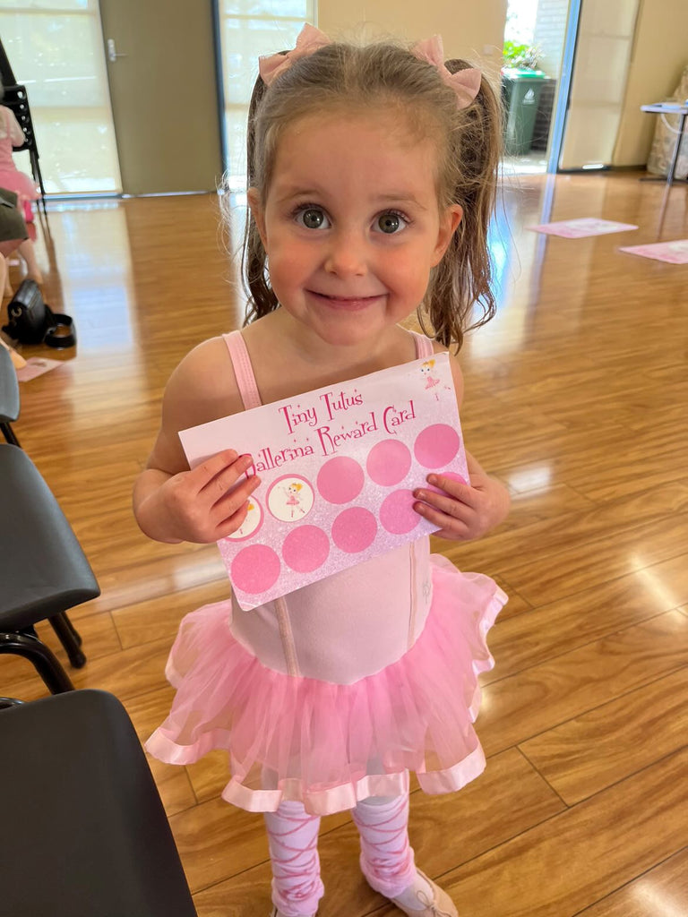 A tiny tutus ballerina in a pink tutu smiling and holding her ballerina reward card with stickers in a ballet class for 3 year olds in Newcastle.