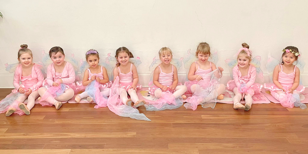 Nine your girls sit together in ballet tutus smiling and playing with dancing scarved in a ballet class for 3 year olds in Adelaide, South Australia.