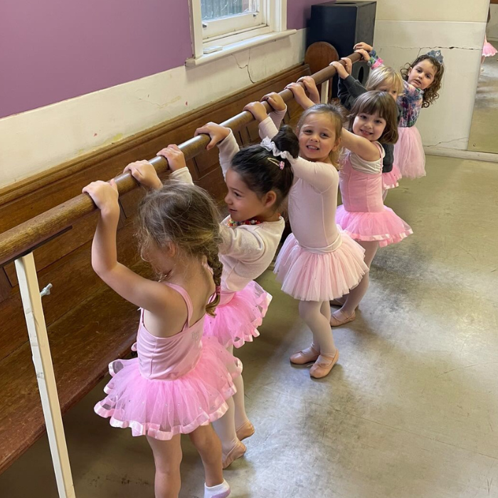 Six young ballerinas stand at a barre in pink tutus on their tippy toes in a ballet class for 5 year olds. 