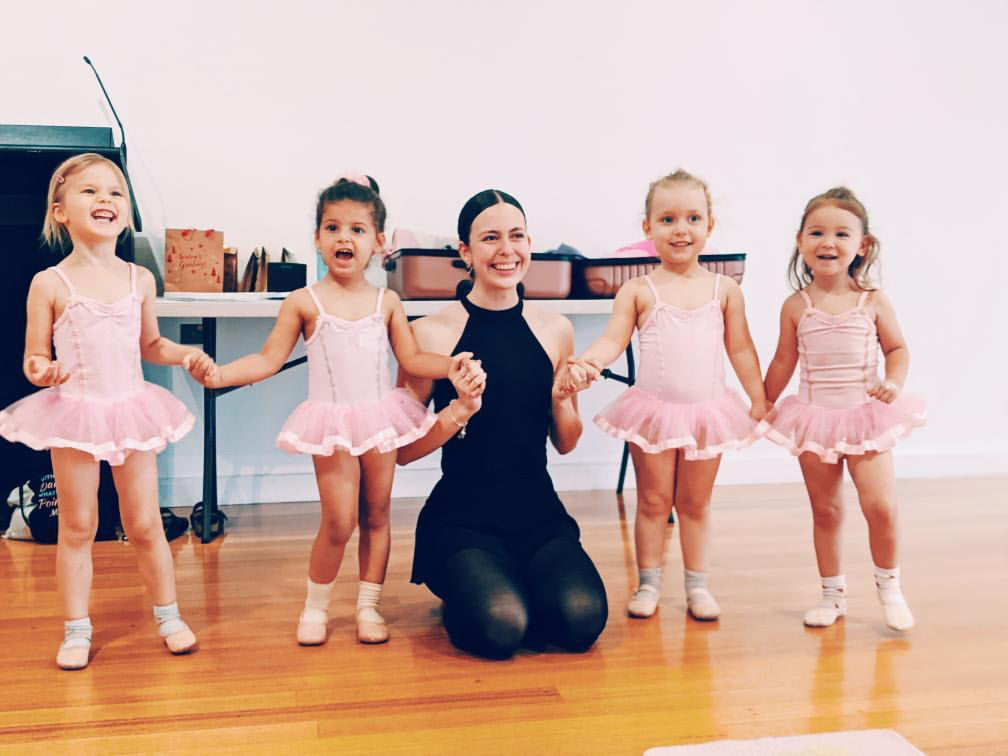 Four ballerinas standing together holding hand with a ballet teacher all wearing pink tutus in a ballet class for 3 year olds. 