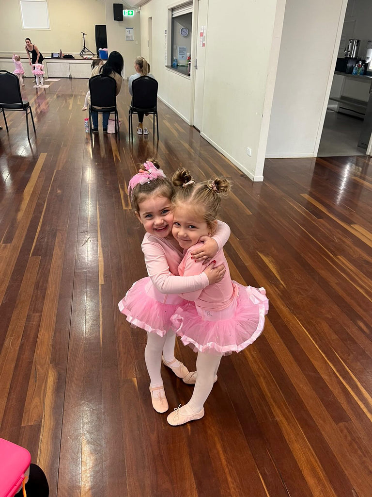 Two ballerinas in pink tutus and jackets hugging and smiling during a ballet class for 3 year olds in Brisbane. 