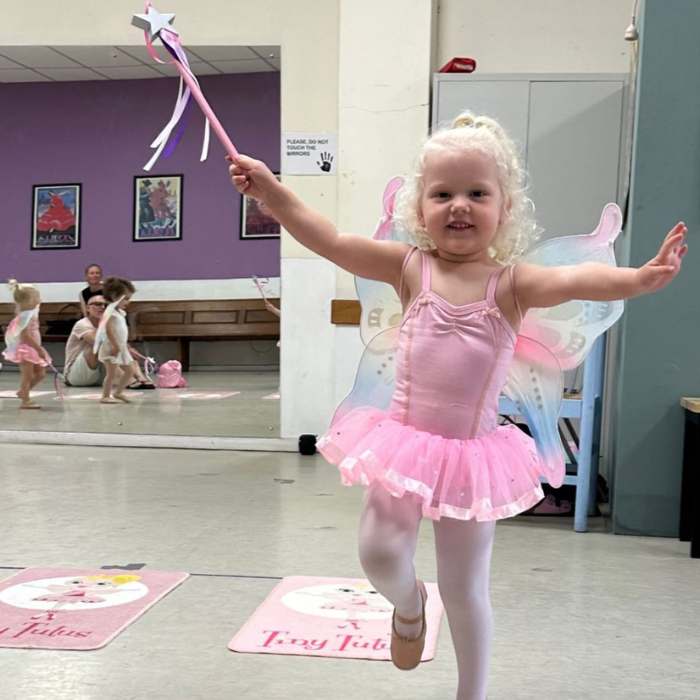 A young ballerina in a pink tutu skips around the classroom wearing fairy wings and waving a fairy wand in a ballet class for 3 year olds. 