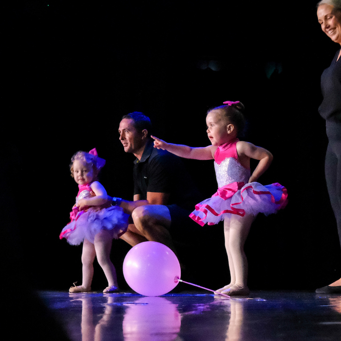 Ballerinas and their parents stand on the stage at their ballet recital with on ballerina pointing to her family in the audience at Newcastle.