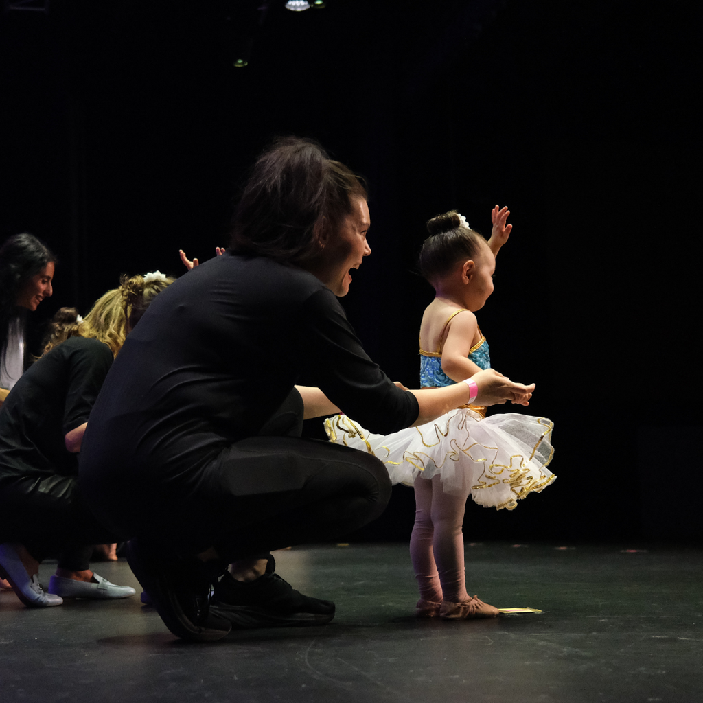 Mum squatting down next to ballerina on stage at a Tiny Tutus recital waving at the audience. 